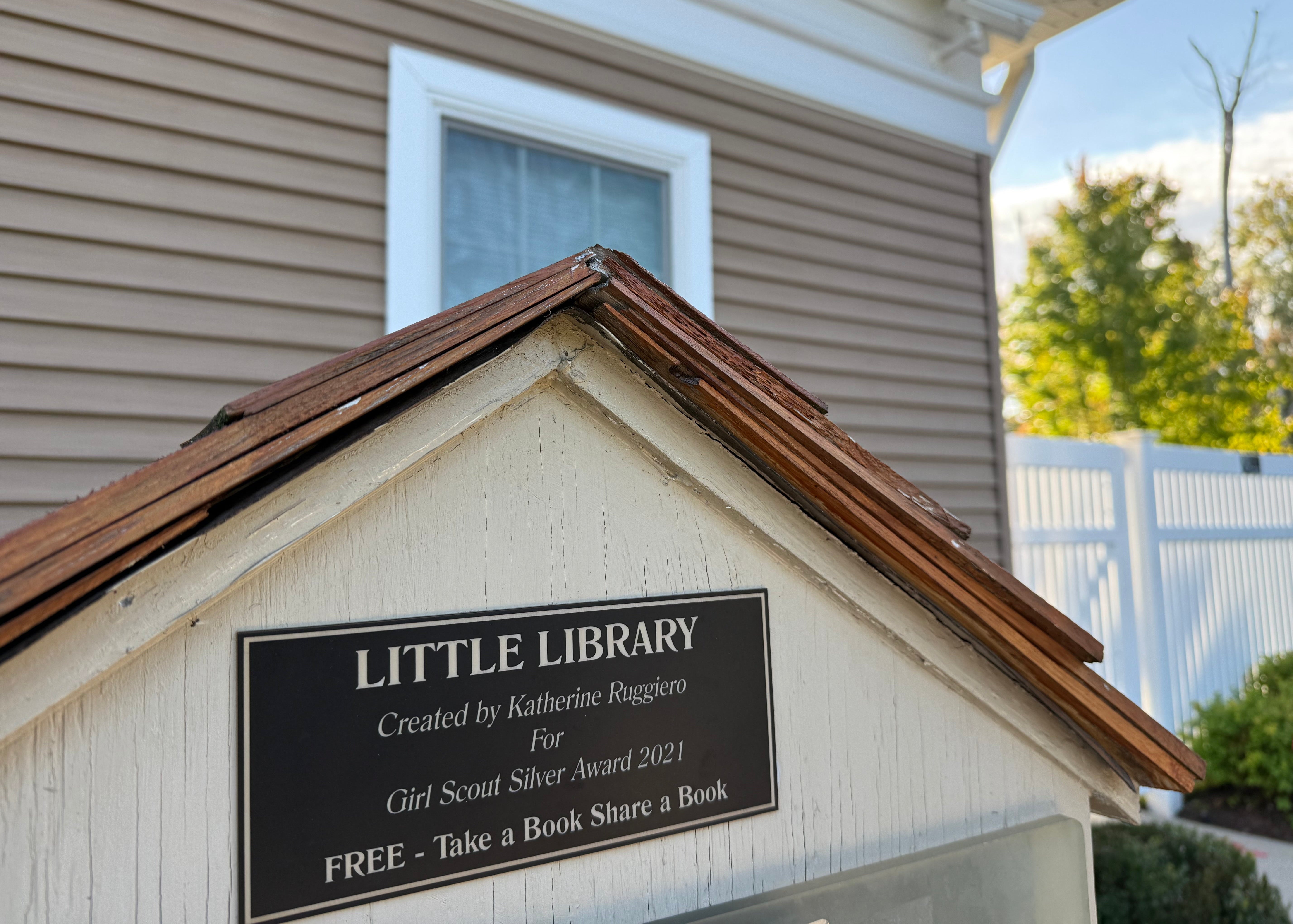 A cream wooden little library with a weathered shingle roof and a black plaque reading "Little Library, Take a Book Share a Book," where The Phoenix Candles donates children's books through Light + Literature.