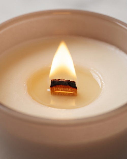 Close-up of the Nurture crystal-infused soy candle by The Phoenix Candles burning inside the matte blush vessel, showing a glowing triangular wooden wick flame above a warm liquid wax pool with the charred wick tip clearly visible, matte blush glass rim framing the composition against a soft white background.