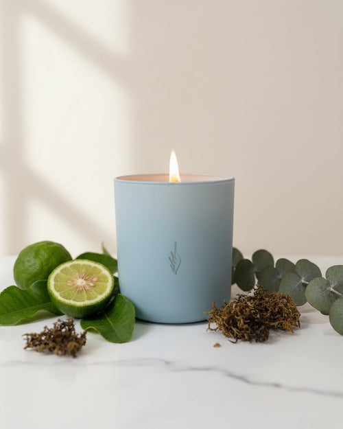 Blue candle with a small logo on a marble surface with limes and eucalyptus leaves.