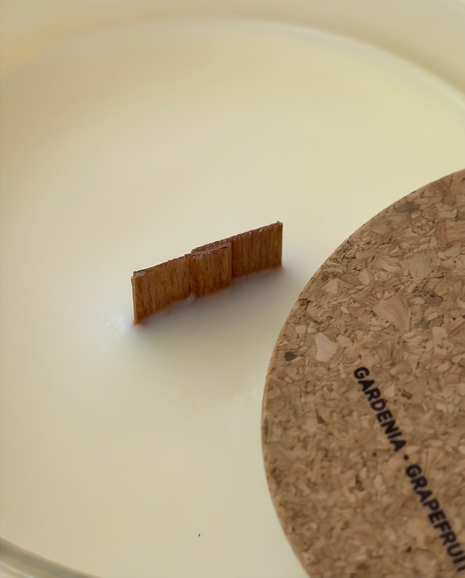 Extreme close-up of the FSC-certified wooden wick centered in the ivory soy wax of The Lab Gardenia and Grapefruit candle by The Phoenix Candles, with the edge of the laser-engraved cork lid reading "Gardenia, Grapefruit" visible in the lower right corner.