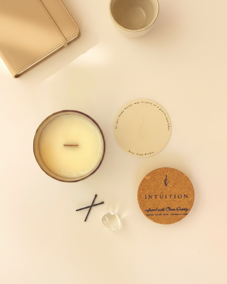 Flat lay of the Intuition crystal-infused soy candle by The Phoenix Candles, featuring an open matte stonewashed grey vessel with wooden wick, cork lid laser-engraved with "Intuition – Infused with Clear Quartz, Casaba Melon, Sage, Gurjum Balsam," a raw Clear Quartz crystal, black match sticks, and a round branded card reading "Rise. From Within.," with a tan leather journal and a speckled ceramic cup partially visible in the upper corners on a clean cream surface.