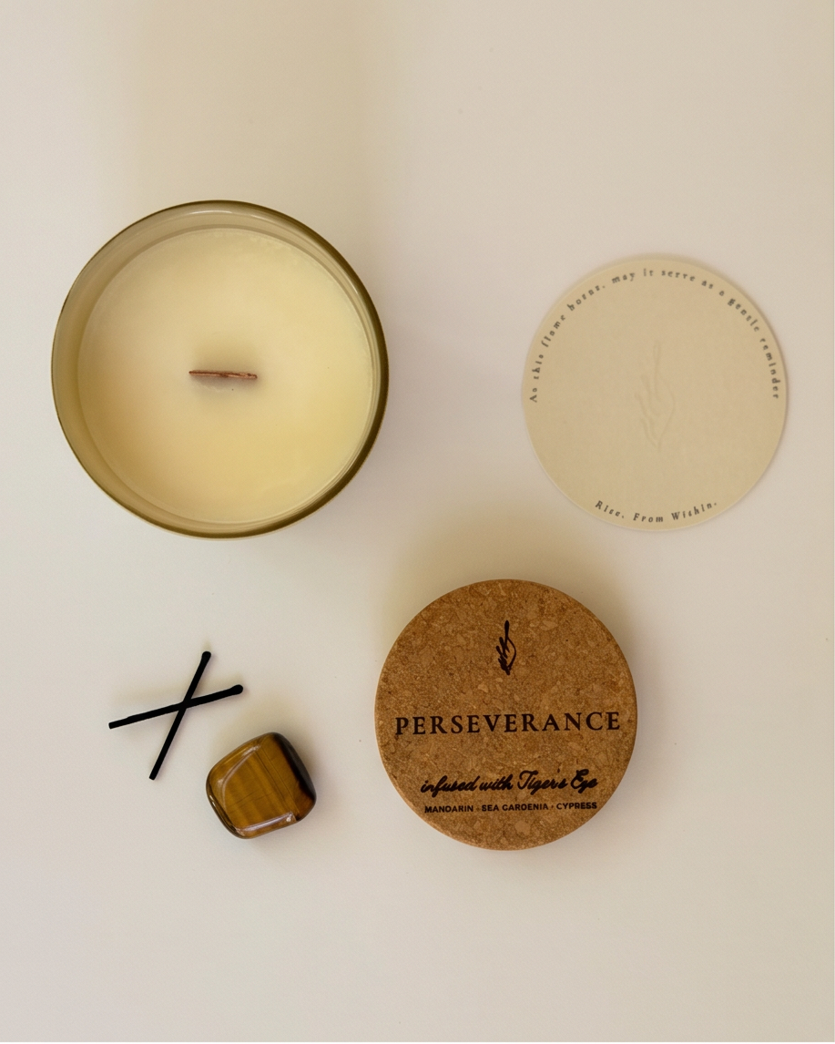 Flat lay of the Perseverance soy candle by The Phoenix Candles, featuring an open matte vessel with wooden wick, cork lid engraved with "Perseverance – Infused with Tiger's Eye, Mandarin, Sea Gardenia, Cypress," a tumbled Tiger's Eye crystal, black match sticks, and a round card reading "Rise. From Within."