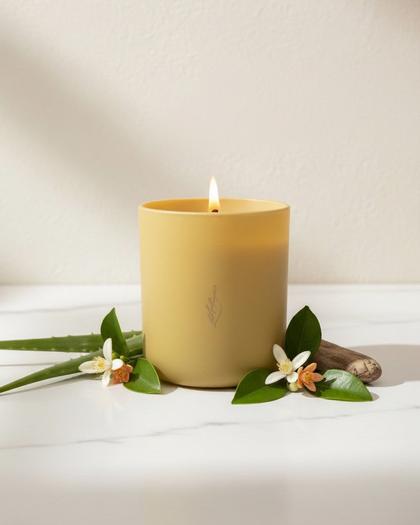 Hope aromachology soy candle by The Phoenix Candles in a matte butter yellow vessel with a lit wooden wick, styled with white orange blossom flowers, fresh aloe vera, and a small piece of driftwood on white marble