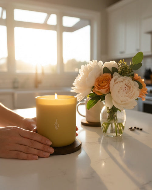 Hands cradling a lit Hope aromachology soy candle in matte yellow vessel on a kitchen counter with flowers