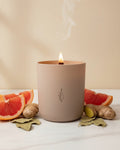Nurture crystal-infused soy candle by The Phoenix Candles in a matte blush vessel, lit wooden wick with rising smoke, surrounded by grapefruit slices, fresh ginger root, and dried bay leaves on white marble.