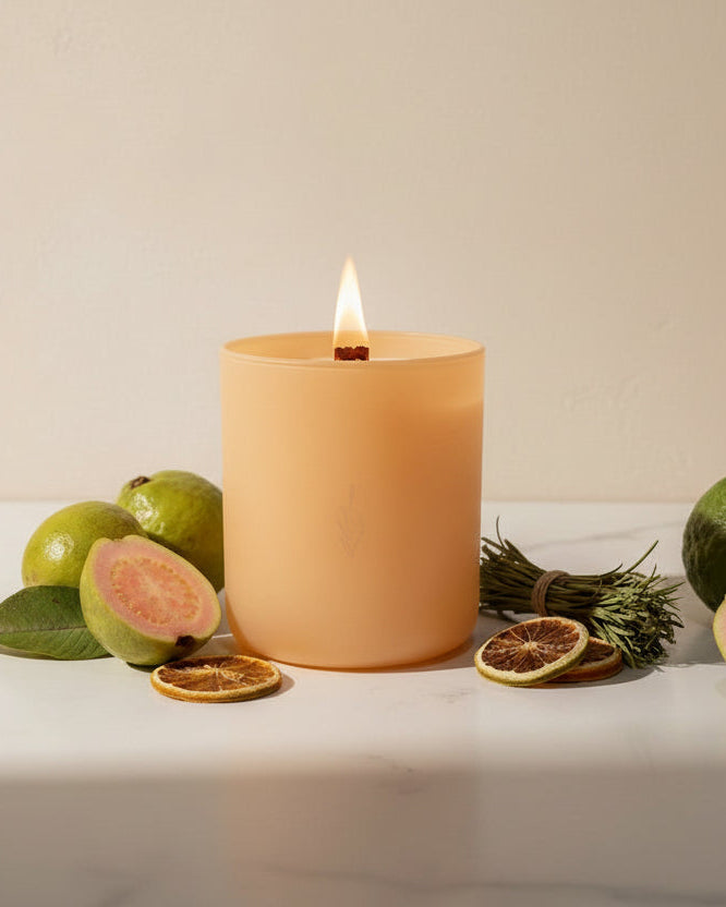 Optimism crystal-infused soy candle by The Phoenix Candles in a matte peach vessel with a lit wooden wick, surrounded by fresh guava fruit, dried blood orange slices, and a bundle of rosemary on white marble.