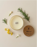 Overhead flat lay of The Lab limited-edition soy candle by The Phoenix Candles, showing an open ivory vessel with wooden wick, a cork lid engraved with "The Lab – Lemon, Rosemary, Jasmine," and fresh scent ingredients including lemon peel, rosemary sprigs, and jasmine blooms arranged on a warm cream background.