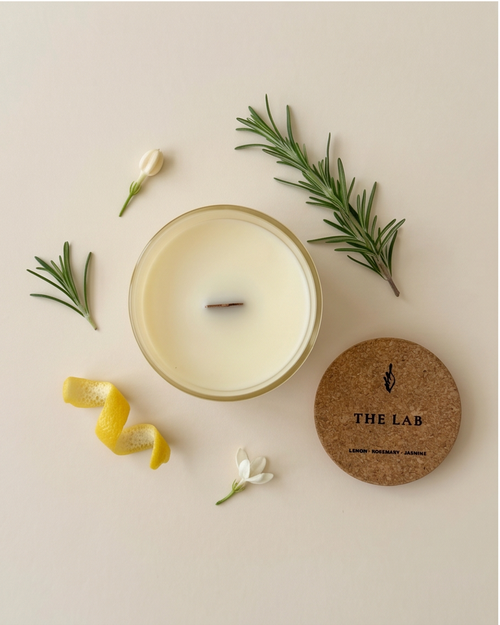 Overhead flat lay of The Lab limited-edition soy candle by The Phoenix Candles, showing an open ivory vessel with wooden wick, a cork lid engraved with "The Lab – Lemon, Rosemary, Jasmine," and fresh scent ingredients including lemon peel, rosemary sprigs, and jasmine blooms arranged on a warm cream background.