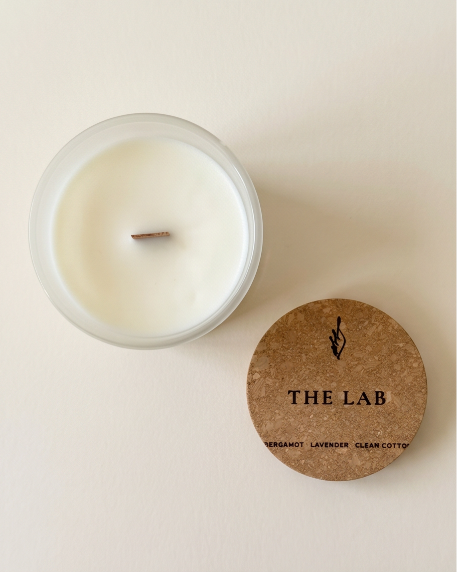 Overhead flat lay of The Lab limited-edition Bergamot, Lavender, and Clean Cotton soy candle by The Phoenix Candles, cork lid removed and placed beside the open frosted clear vessel revealing a smooth ivory wax pour and FSC-certified wooden wick, lid laser-engraved with "The Lab – Bergamot, Lavender, Clean Cotton" on a warm cream background.