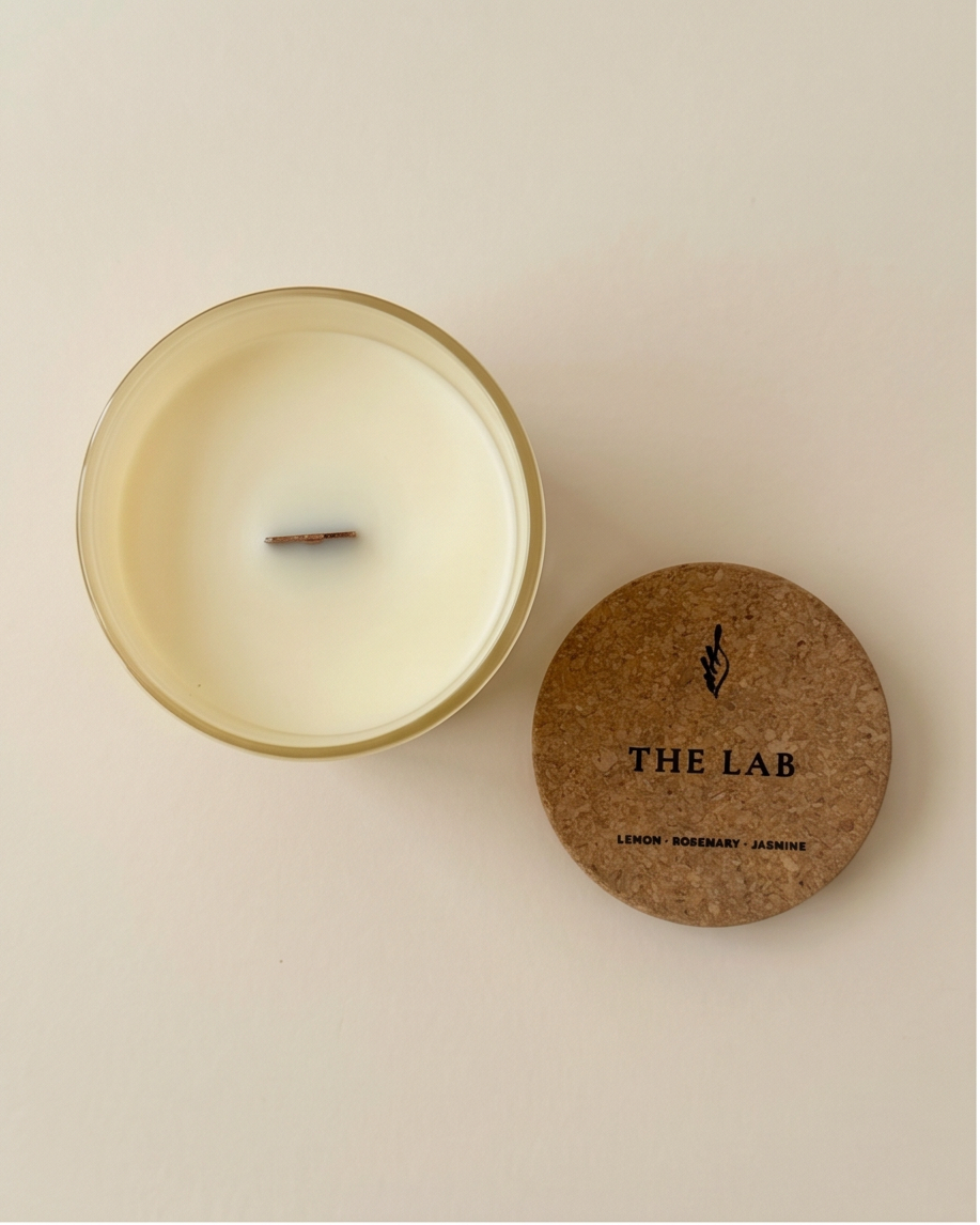 Overhead flat lay of The Lab limited-edition soy candle by The Phoenix Candles, with the cork lid removed and placed beside the open frosted clear vessel, revealing smooth ivory soy wax and an FSC-certified wooden wick, on a warm cream background.