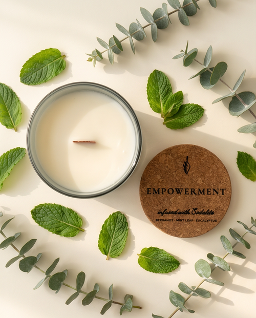 Overhead flat lay of the Empowerment crystal-infused soy candle by The Phoenix Candles, open matte cloud blue reusable glass vessel with wooden wick centered, cork lid laser-engraved with "Empowerment – Infused with Sodalite, Bergamot, Mint Leaf, Eucalyptus" placed to the lower right, surrounded by fresh mint leaves and silver eucalyptus sprigs scattered abundantly on a warm cream background.