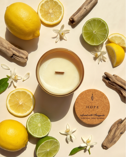Overhead flat lay of the Hope crystal-infused soy candle by The Phoenix Candles, open matte canary yellow vessel with wooden wick centered, cork lid laser-engraved with "Hope – Infused with Amazonite, Blanc Citrus, Pcholi, Driftwood" placed to the lower right, surrounded by whole and halved lemons, halved limes, small white citrus blossoms, and pieces of driftwood on a warm cream background in strong natural light.