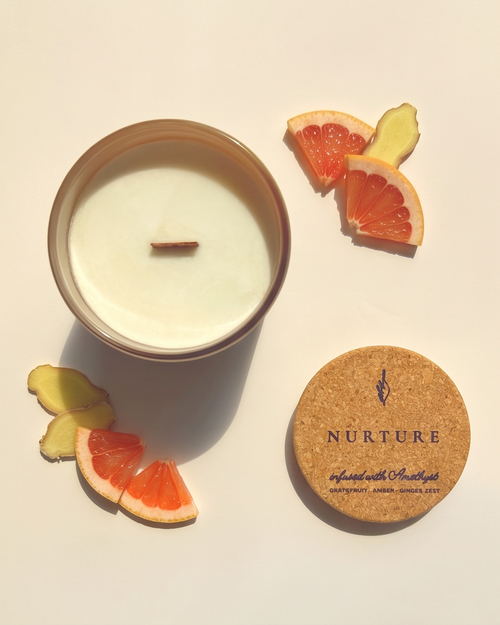 Overhead flat lay of the Nurture crystal-infused soy candle by The Phoenix Candles, open matte blush reusable glass vessel with wooden wick centered, cork lid laser-engraved with "Nurture – Infused with Amethyst, Grapefruit, Amber, Ginger Zest" placed to the lower right, surrounded by fresh grapefruit wedges and sliced ginger root on a warm cream background with dramatic natural light and shadow.