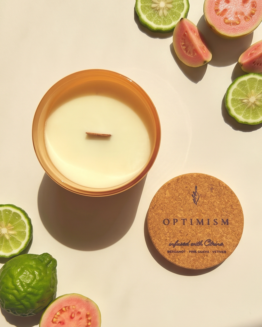 Overhead flat lay of the Optimism crystal-infused soy candle by The Phoenix Candles, open matte peach reusable glass vessel with wooden wick centered, cork lid laser-engraved with "Optimism – Infused with Citrine, Bergamot, Pink Guava, Vetiver" placed to the lower right, surrounded by halved pink guava, sliced limes, and whole bergamot fruit on a warm cream background in dramatic natural light and shadow.