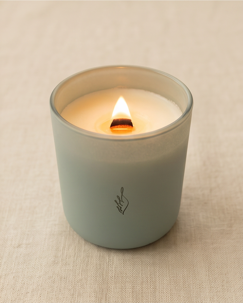 The Empowerment crystal-infused soy candle by The Phoenix Candles burning in a matte cloud blue reusable glass vessel with laser-engraved brand flame logo and wooden wick flame glowing warm amber, displayed lidless on a natural woven linen surface.