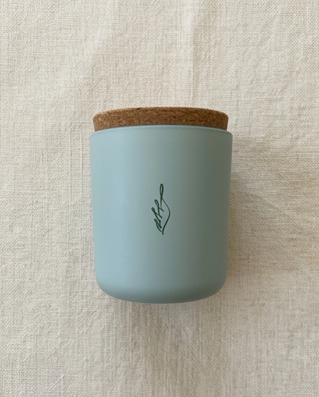 The Empowerment crystal-infused soy candle by The Phoenix Candles in a matte cloud blue reusable glass vessel with natural cork lid and laser-engraved brand flame logo centered on the front, photographed straight-on against a natural woven linen textile background.