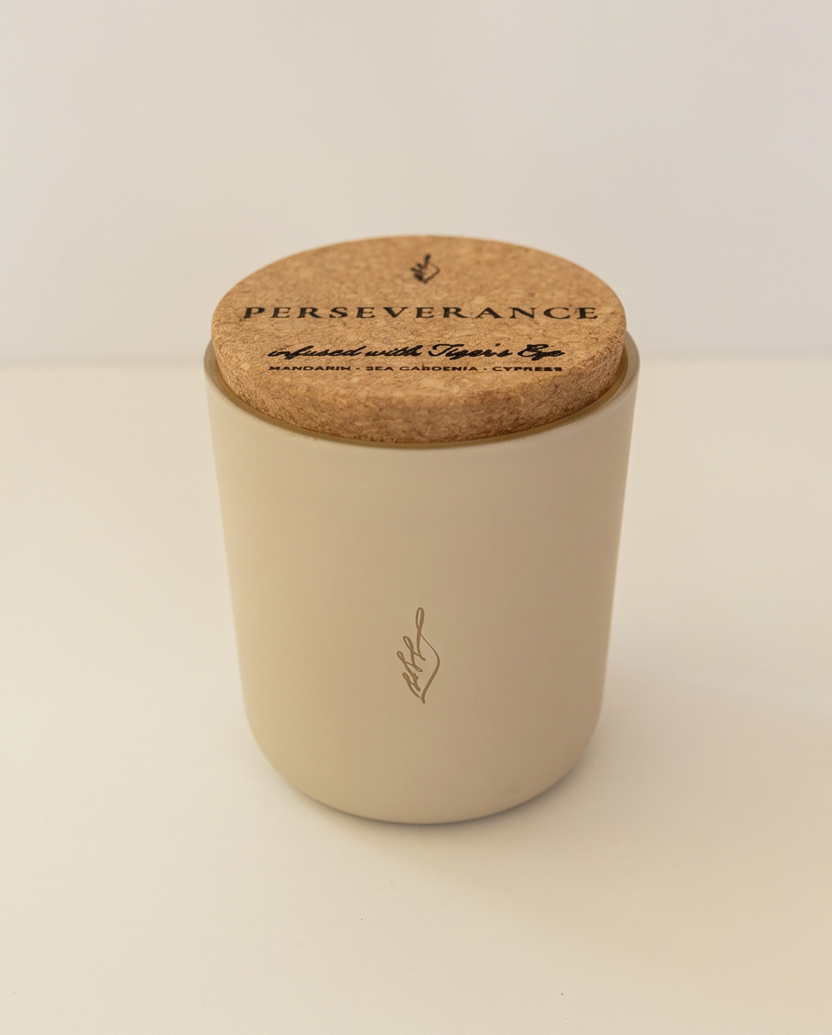 The Perseverance soy candle by The Phoenix Candles in a matte cream reusable glass vessel with a natural cork lid laser-engraved with "Perseverance – Infused with Tiger's Eye, Mandarin, Sea Gardenia, Cypress," and a subtle flame logo embossed on the front, photographed against a clean white background.