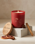 The Year of the Fire Horse limited-edition soy candle by The Phoenix Candles burning in a matte red reusable glass vessel with a laser-engraved rearing horse illustration, displayed on a concrete pedestal with the cork lid laser-engraved "Year of the Fire Horse – Infused with Fire Opal, Blood Orange, Spices, Cedarwood" resting beside it, styled with dried blood orange slices, whole star anise, and a cedarwood stick on a warm cream surface.
