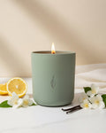Xenacious aromachology soy candle by The Phoenix Candles in a matte sage green vessel with a lit wooden wick, styled with halved lemons, white jasmine blossoms, and vanilla bean pods on white marble