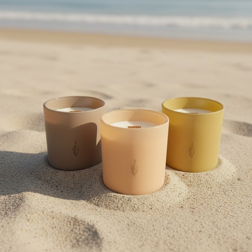The Phoenix Candles Summer Edit, three soy candles in blush, peach, and yellow vessels on a sandy beach with the ocean behind.