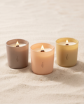 The Phoenix Candles Summer Edit bundle, three lit soy candles in blush, peach, and butter-yellow vessels grouped on pale beach sand.